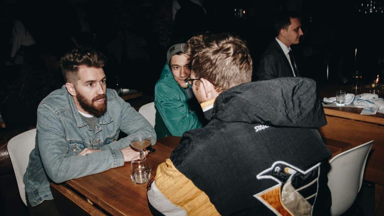 Three men are seated at a table, looking at a fourth man in a black and yellow jacket.