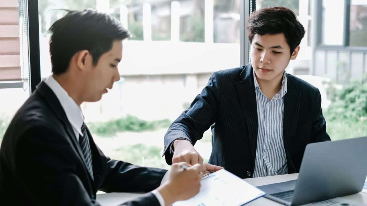 Two men in business suits are sitting at a desk with a laptop, looking at a document.