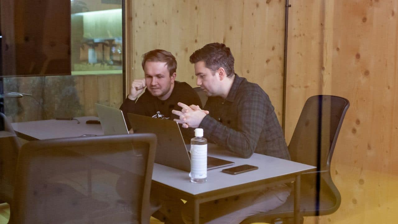 Two men are sitting at a table in an office, looking at a laptop.