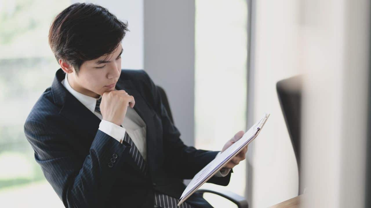 A young man in a suit is sitting in a chair, holding a clipboard and looking at a document.