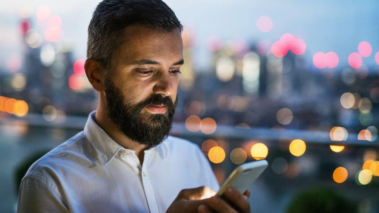 A man with a beard is looking at his phone at night with city lights in the background.