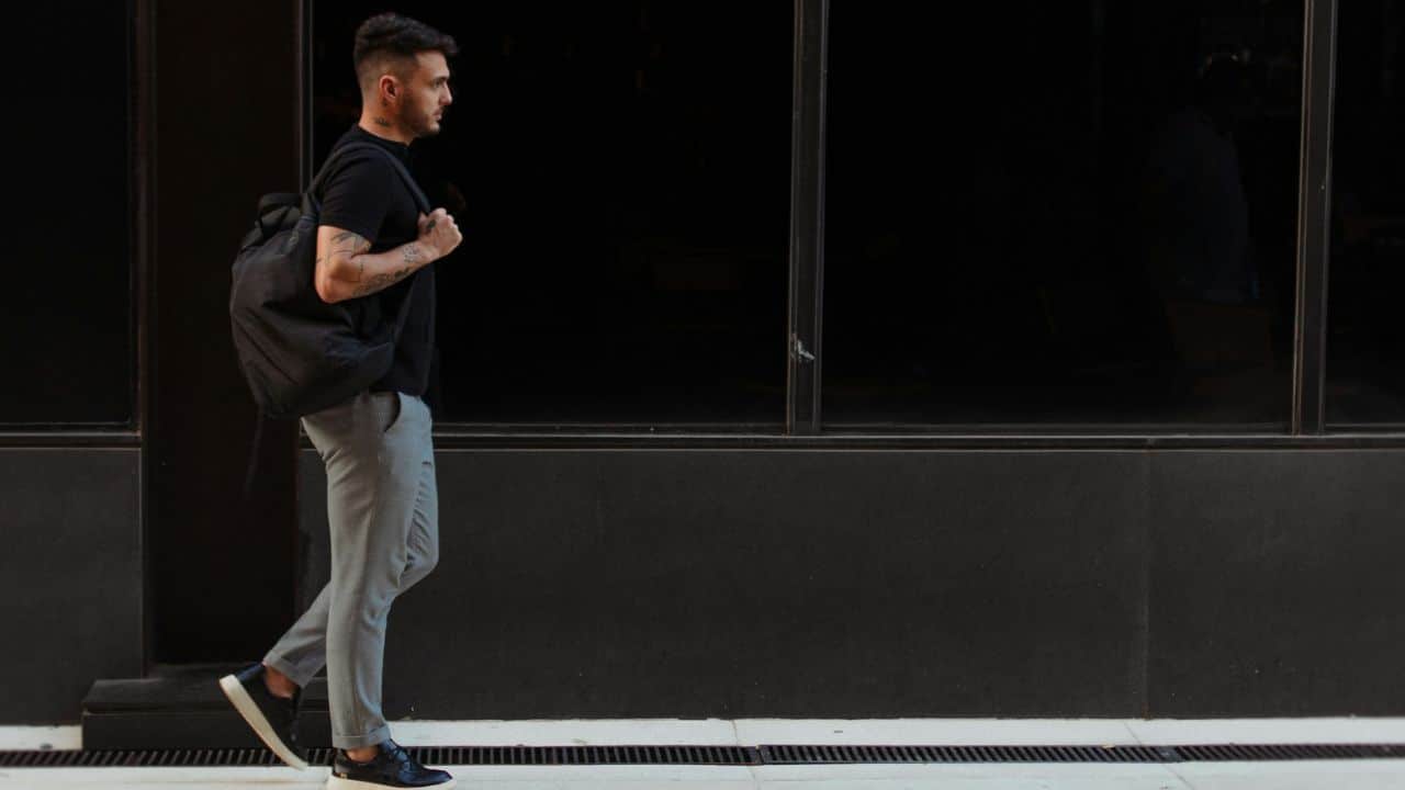 A man with tattoos on his arms is walking on a sidewalk, carrying a black backpack.