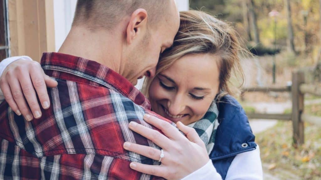 A smiling man in a red and black plaid shirt hugs a woman with her eyes closed.