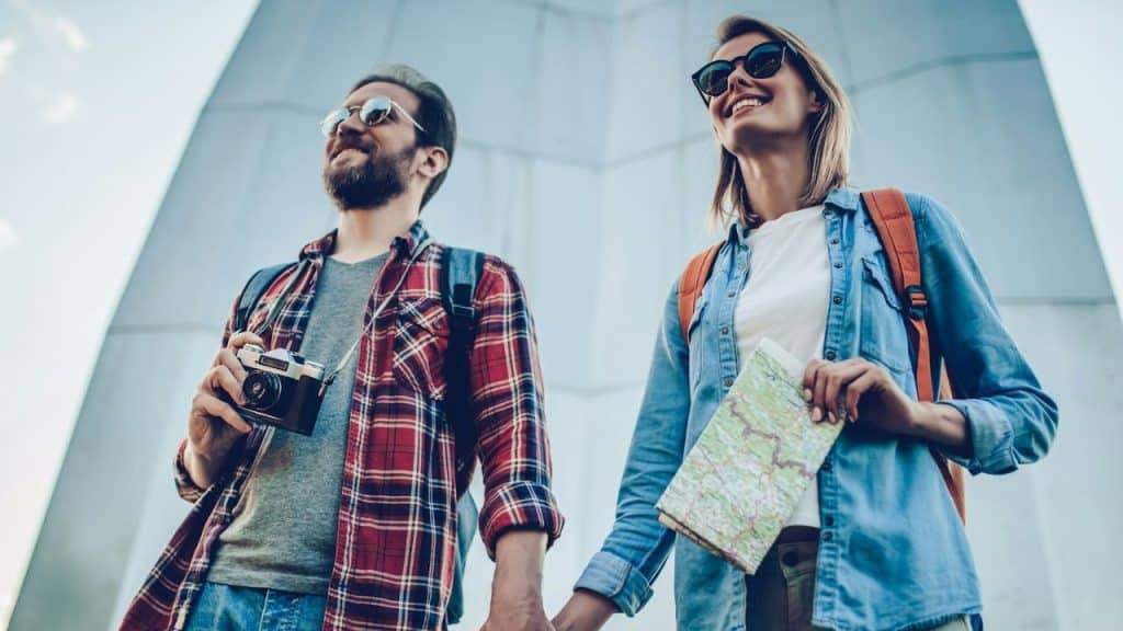 A happy, young couple holding hands and wearing sunglasses and backpacks stands outside.