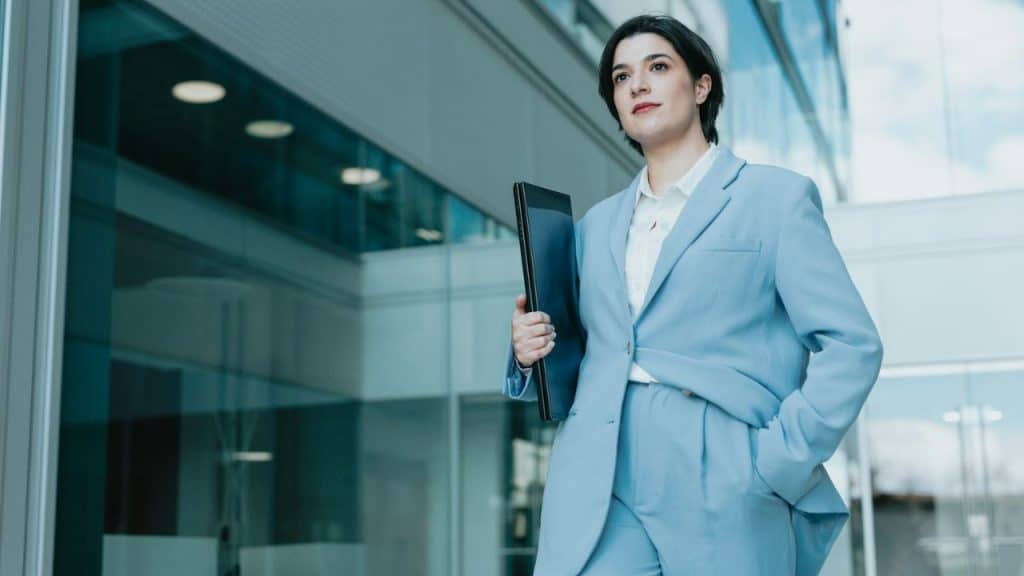 A smiling, confident businesswoman in a light blue suit stands in front of a modern building, holding a black folder.
