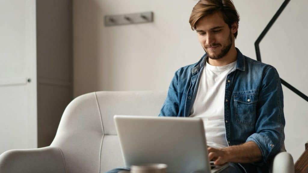 A happy man with a beard sits on a couch, looking at a laptop.