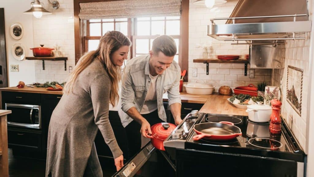 A smiling couple looks into an oven, holding a red pot together.