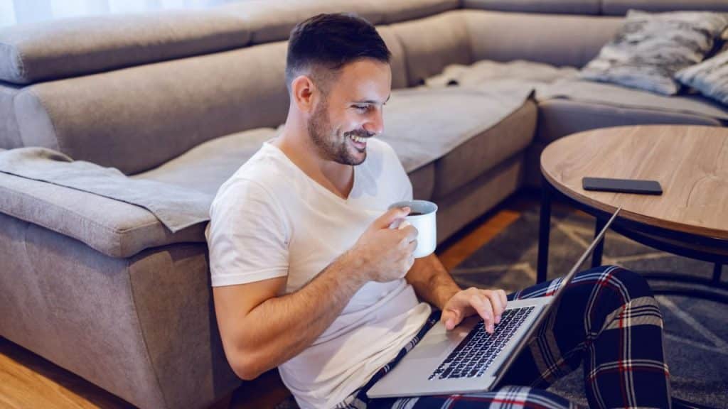A happy man sits on the floor with a laptop, drinking from a mug.