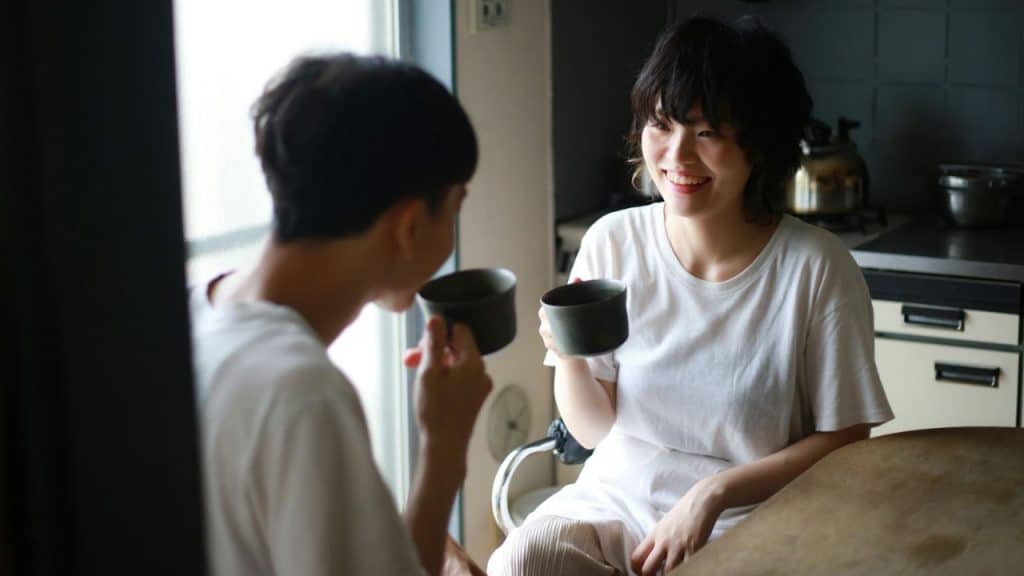 A happy couple sits at a kitchen table, holding mugs and looking at each other.