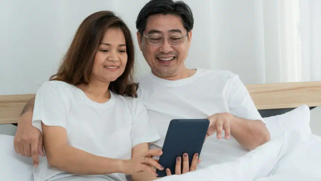A happy couple smiles in bed, looking at a tablet together.