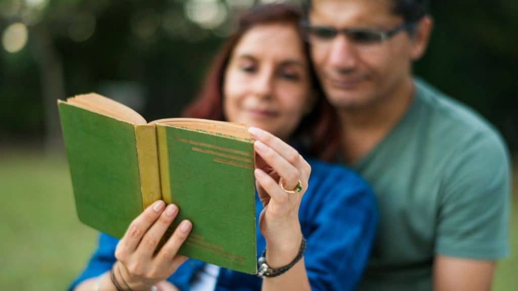 A woman and a man sit together outdoors, reading an open green book.