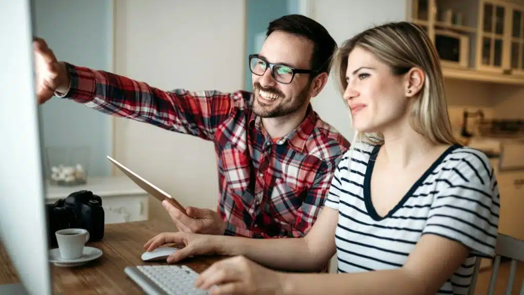 A smiling couple works at a desk, looking at a computer monitor and a tablet.