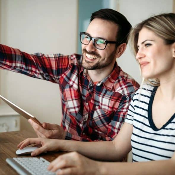 A smiling couple works at a desk, looking at a computer monitor and a tablet.