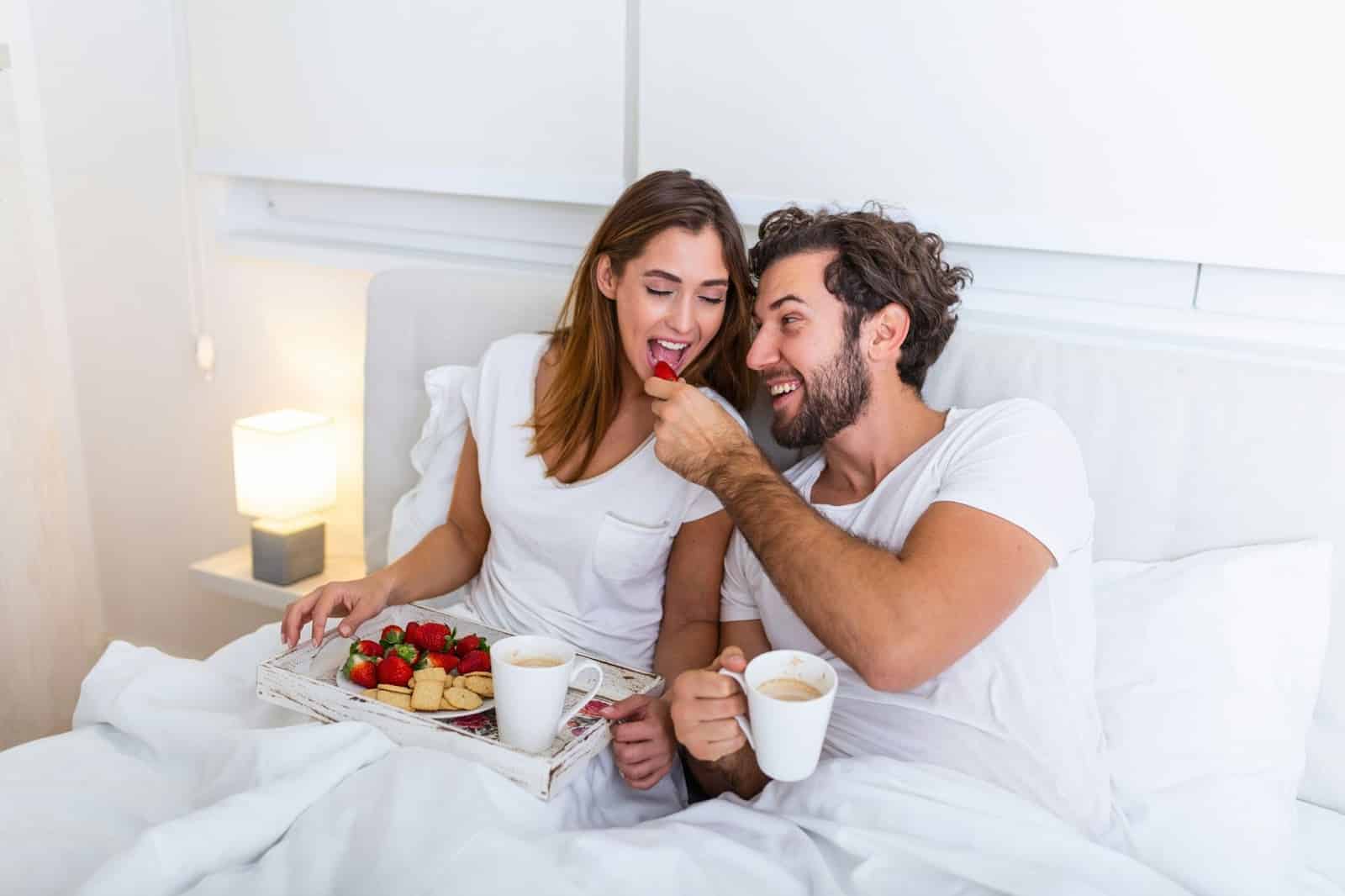 A man and woman eating breakfast at the bed.