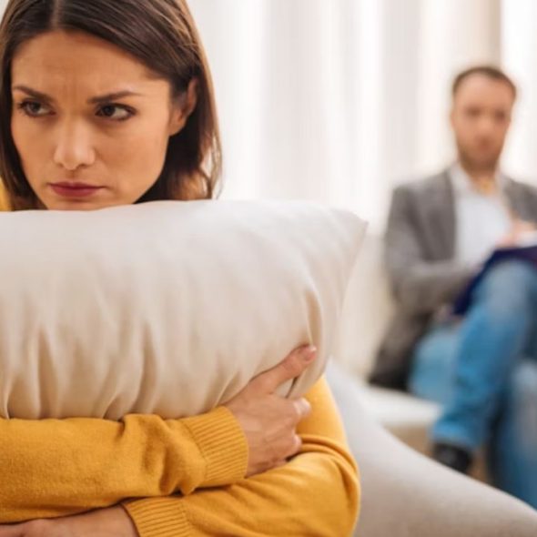 A woman with a sad expression sits on a couch, holding a pillow, with a man blurred in the background.
