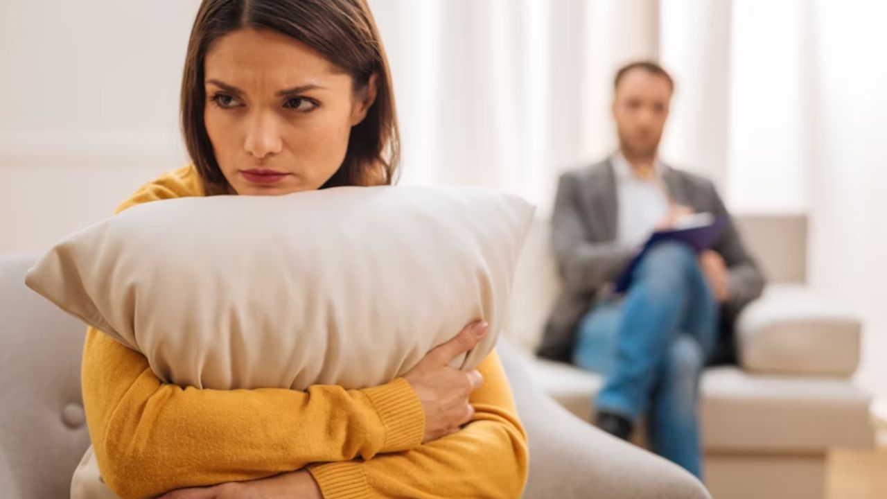 A woman with a sad expression sits on a couch, holding a pillow, with a man blurred in the background.