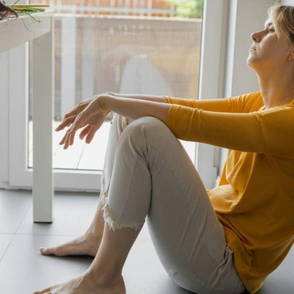 A woman in a yellow shirt and light-colored pants is sitting on the floor, leaning against a wall.
