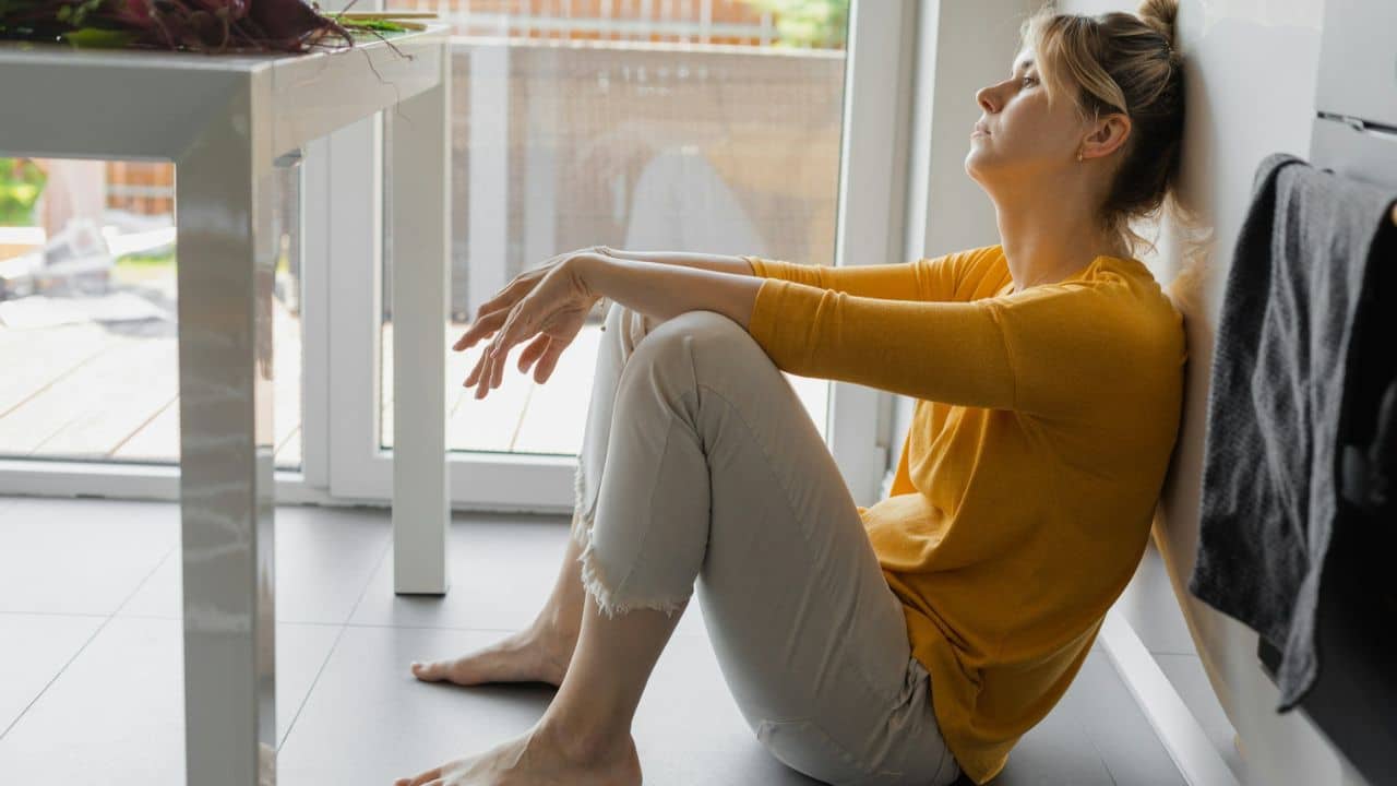 A woman in a yellow shirt and light-colored pants is sitting on the floor, leaning against a wall.