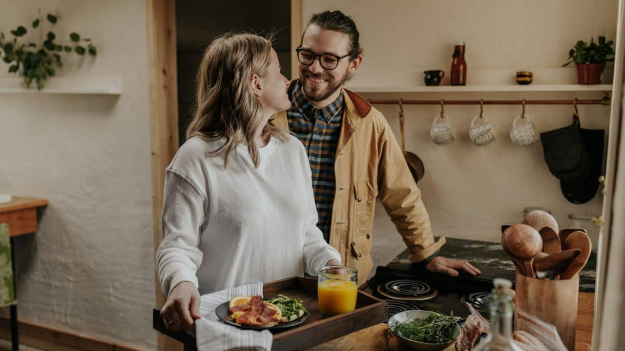 A smiling man is watching a woman in a kitchen as she holds a tray with a drink and food.