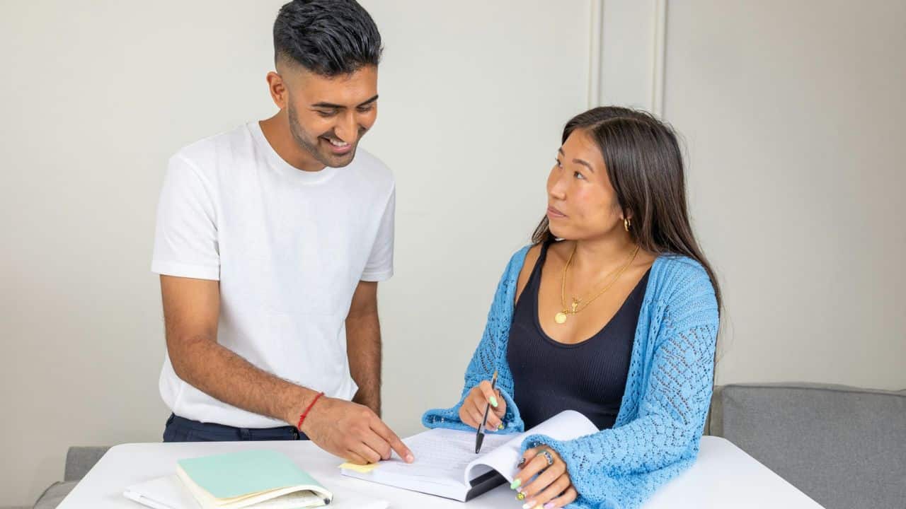A smiling man is standing next to a woman who is sitting at a table and writing in a notebook.
