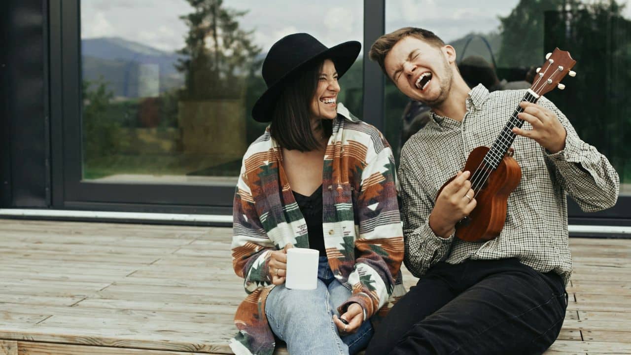 A couple is laughing together on a wooden deck, and the man is playing a ukulele.