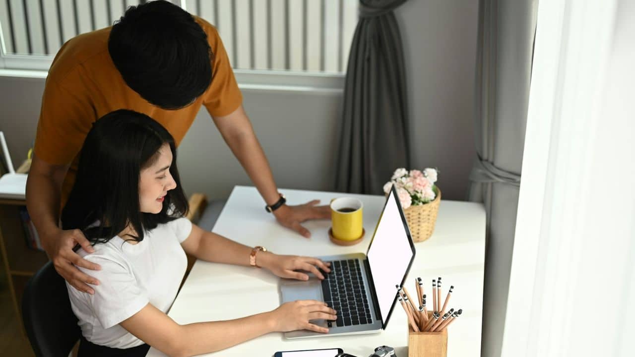 A man is standing behind a woman who is sitting at a desk and using a laptop.
