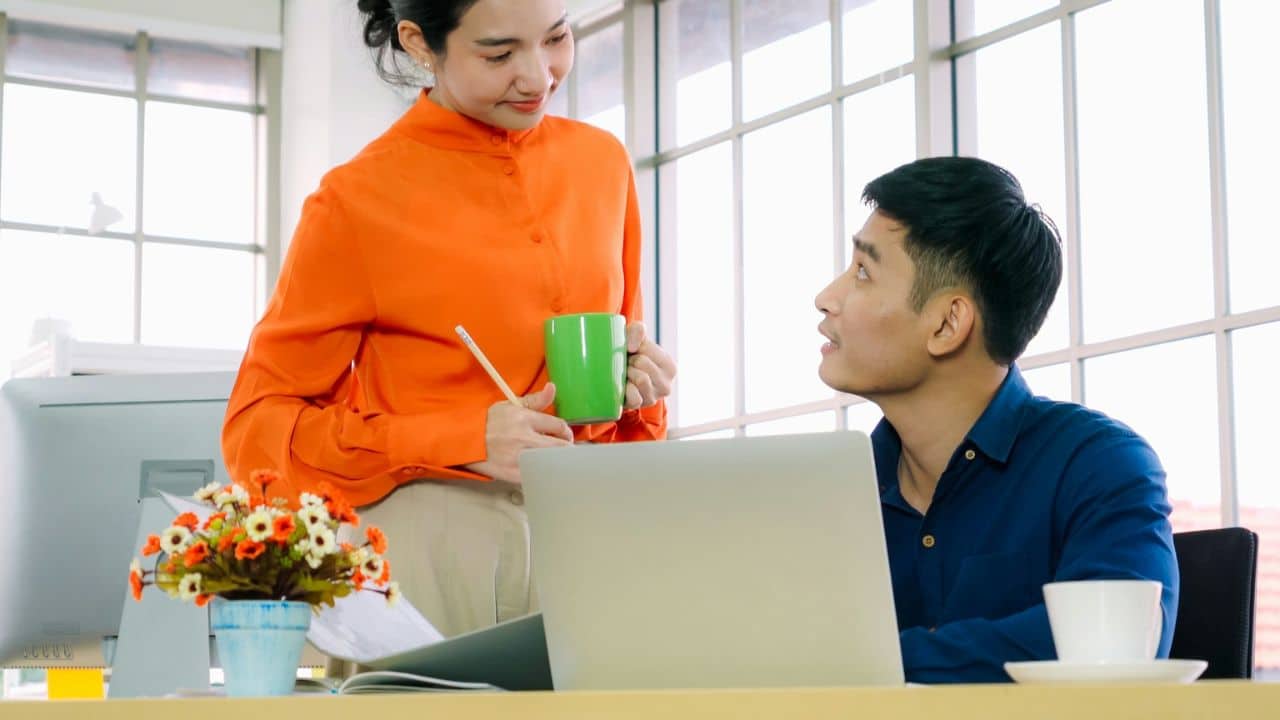 A woman in an orange shirt is standing and talking to a man who is sitting at a desk.