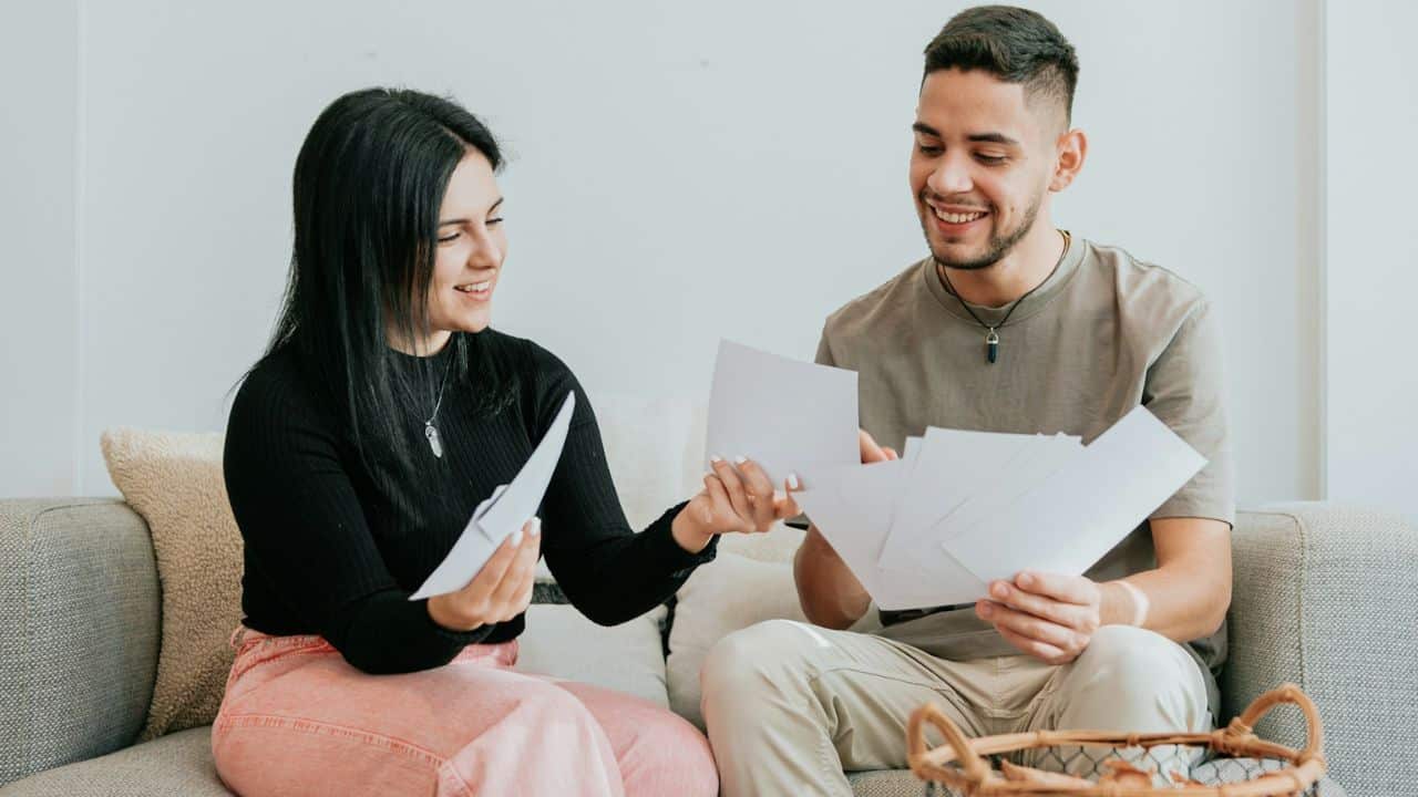 A smiling couple is sitting on a couch and looking at papers together.