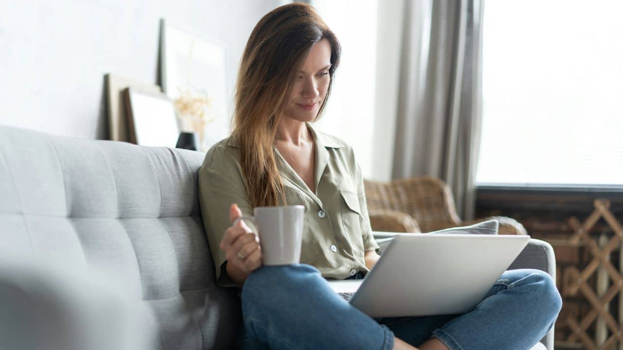 A woman is sitting on a gray couch, holding a mug and looking at a laptop.