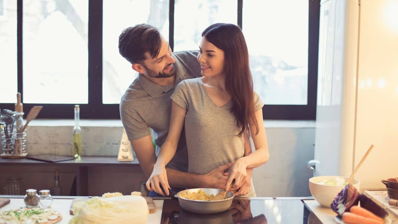 A smiling couple is in a kitchen, looking at each other while a man stands behind a woman with his arms around her.