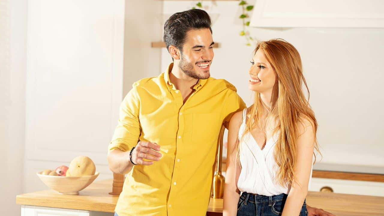 A smiling couple is in a kitchen, looking at each other while a man leans on a countertop.