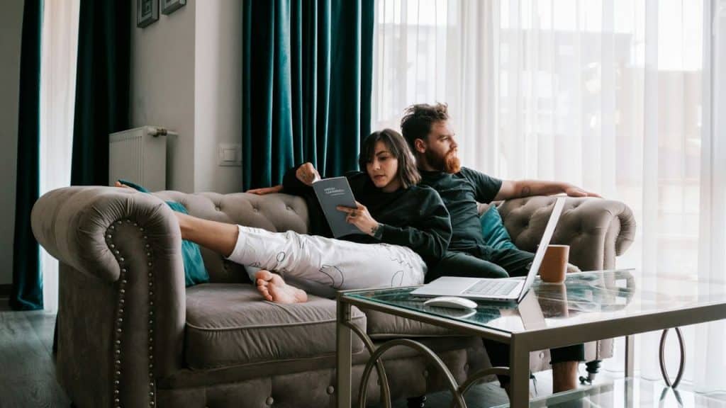 A woman reads a book while a man looks out the window, sitting on a couch.