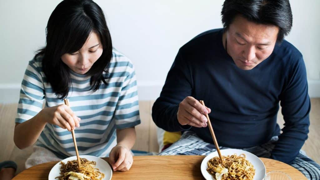 A couple sits at a low table, eating noodles from plates with chopsticks.
