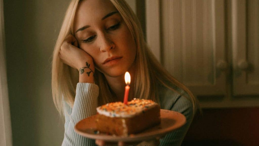 A sad woman with a tattoo on her arm looks at a slice of cake with a lit candle.