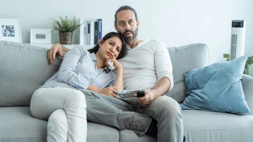 A sad woman leans on a man's shoulder while he looks surprised while holding a remote.