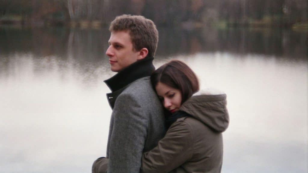 A woman with a sad expression hugs a man from behind, resting her head on his back. They are standing near a body of water.