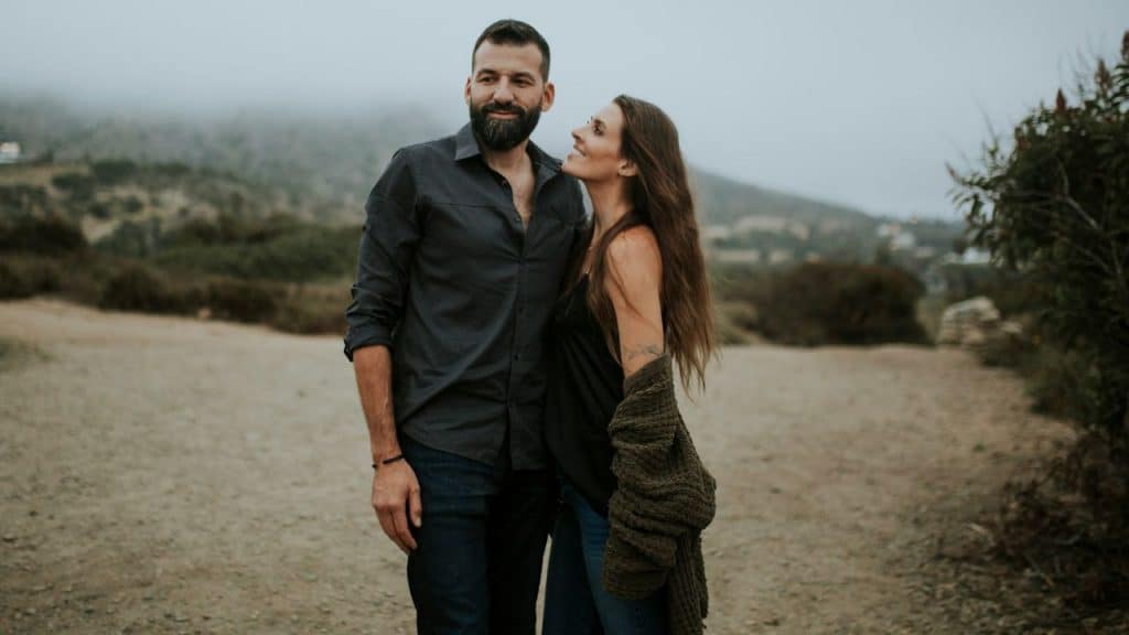 A smiling couple looks at each other while standing on a dirt road in a field.