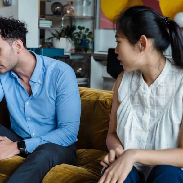 A man is sitting on a couch with his head in his hand, and a woman is sitting nearby, talking to him.