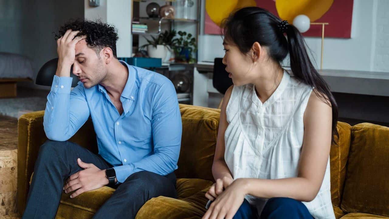 A man is sitting on a couch with his head in his hand, and a woman is sitting nearby, talking to him.