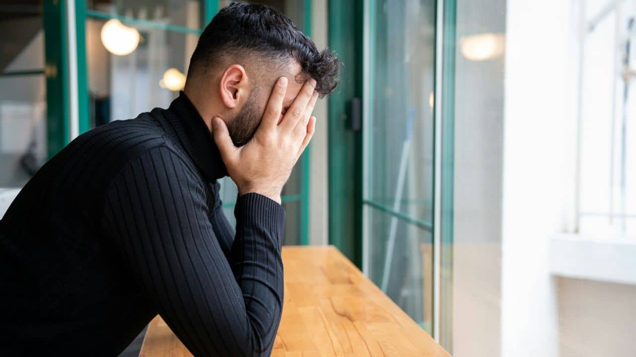 A man is sitting at a table with his face in his hands, looking stressed.