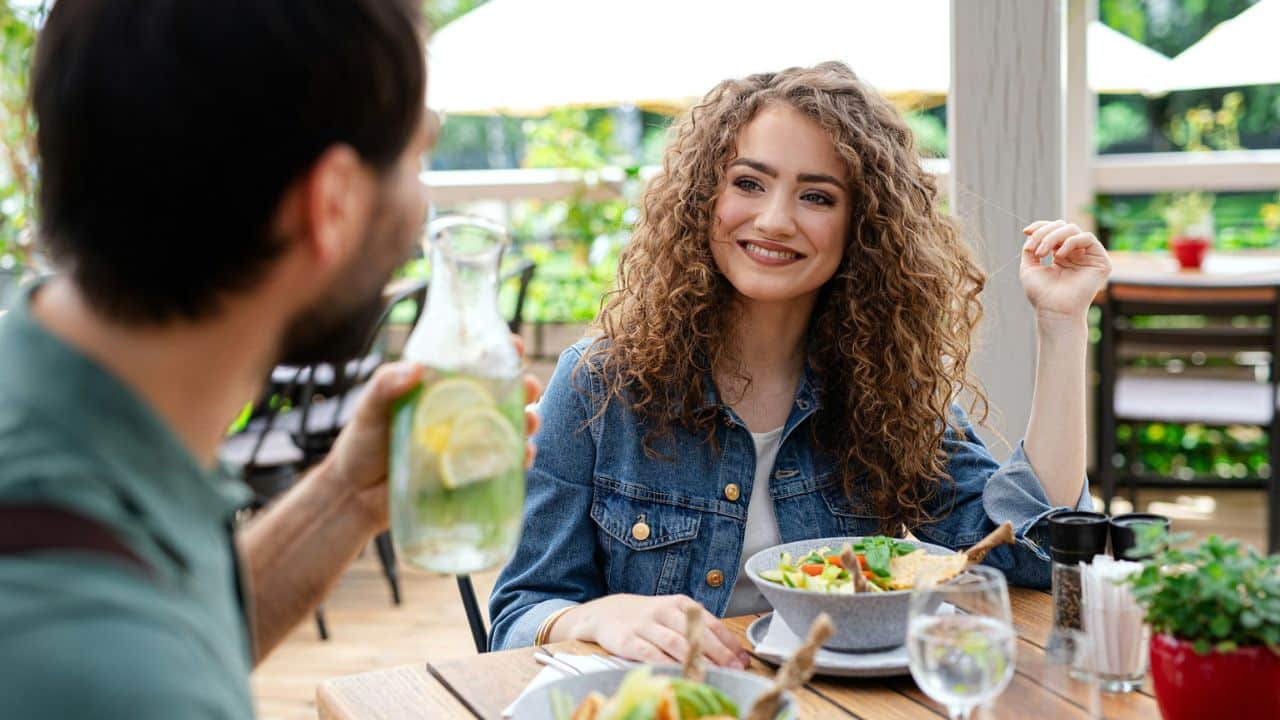 A smiling woman with curly hair is sitting across from a man at a table, with food and drinks in front of them.