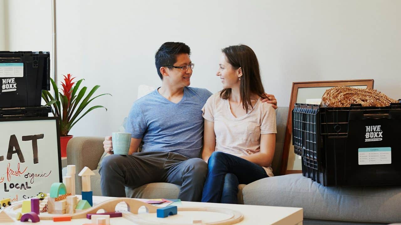 A couple is sitting on a couch, smiling at each other, with moving boxes on either side of them.