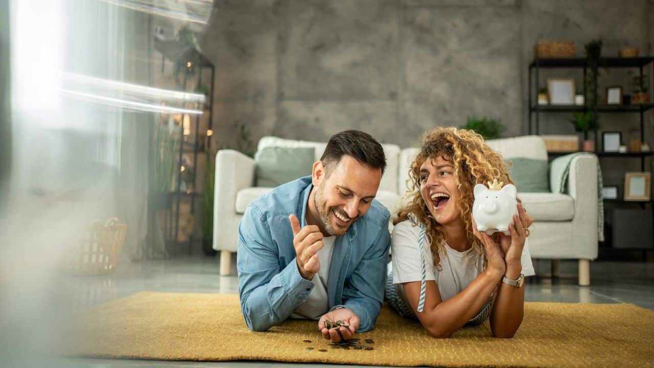 A couple lying on the floor, smiling while holding money and a piggy bank.