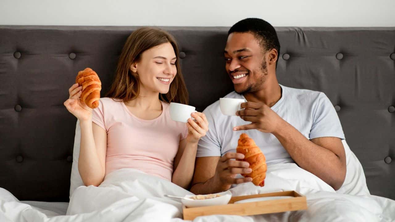 A man and woman smiling at each other while having croissants and coffee in bed.