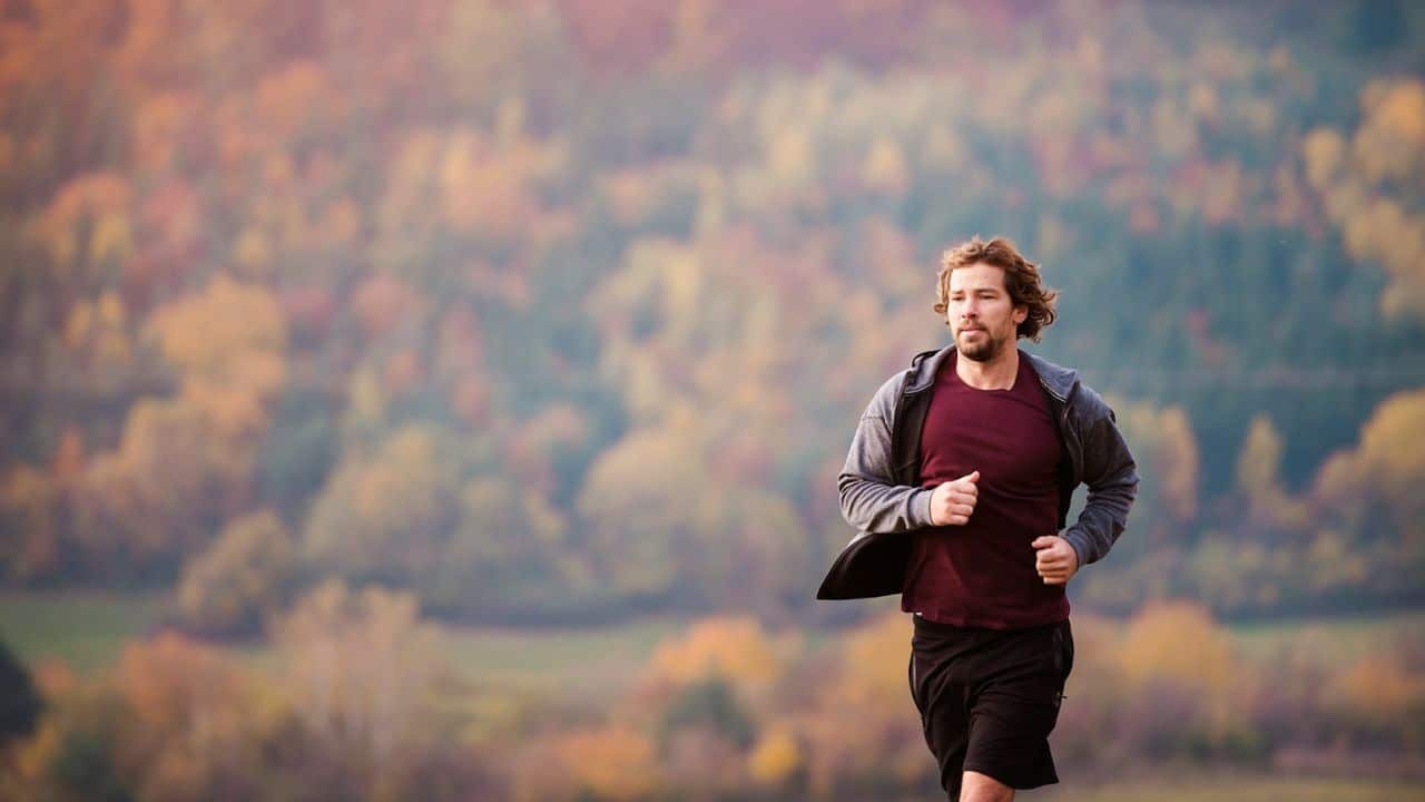 A man with a beard running outdoors in front of fall-colored trees.