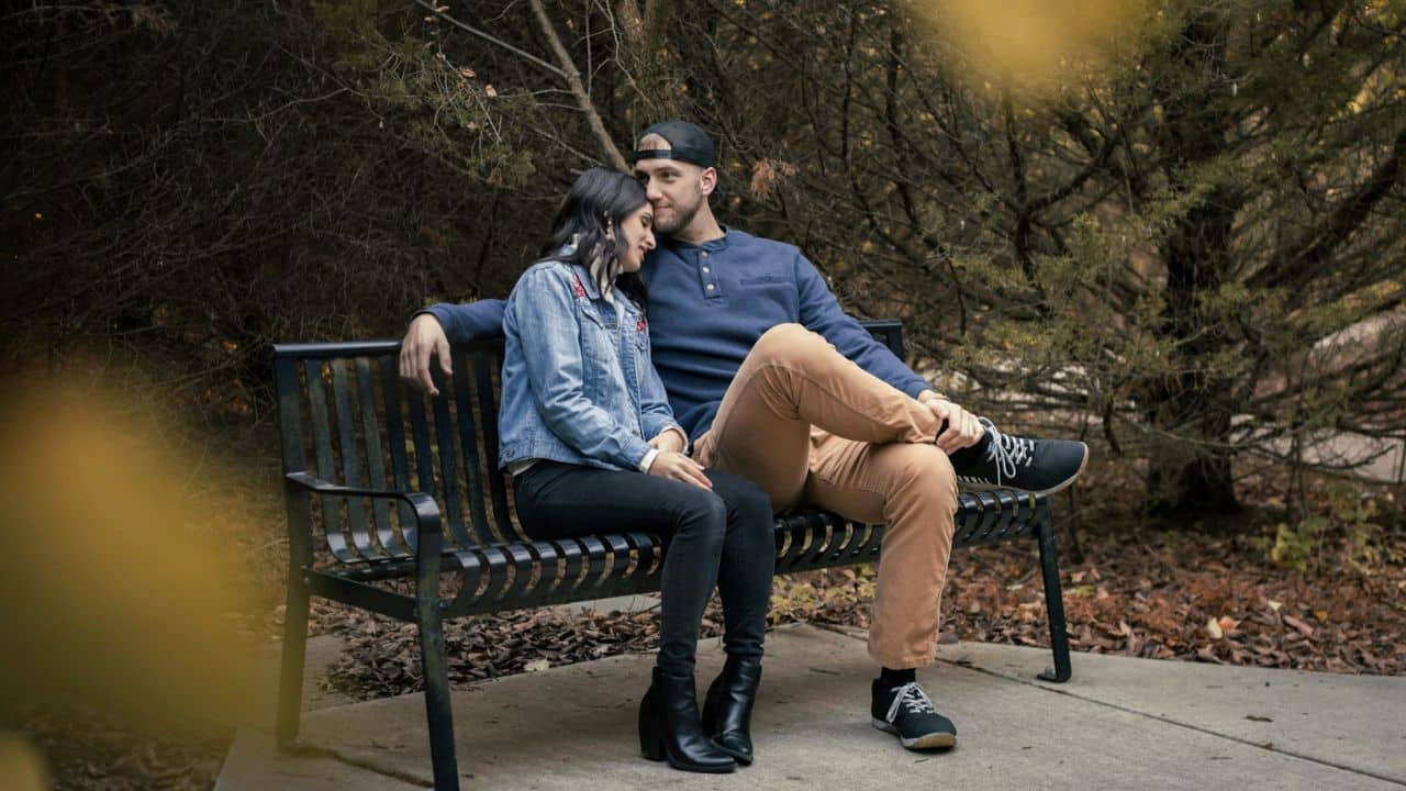 A man and a woman sitting on a park bench under a tree.