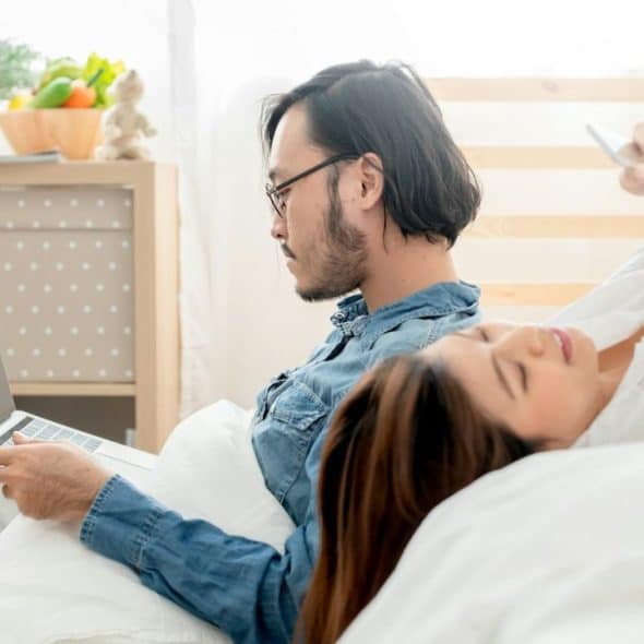 A couple in bed looking at their phones and a laptop.