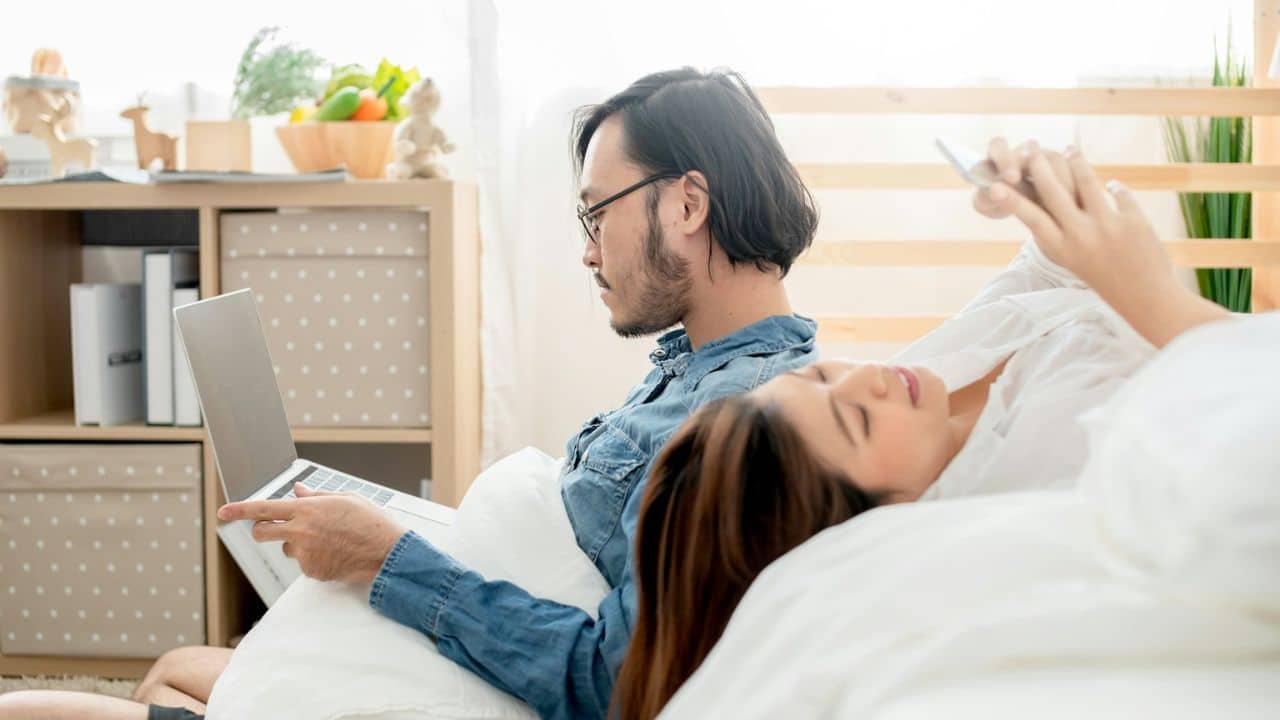A couple in bed looking at their phones and a laptop.