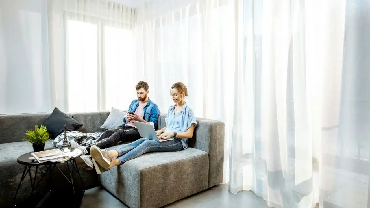 A man and woman sit on a sofa, looking at their separate phones and a laptop.