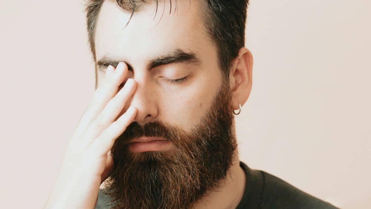 A tired man with a beard and earring rubbing his eyes.